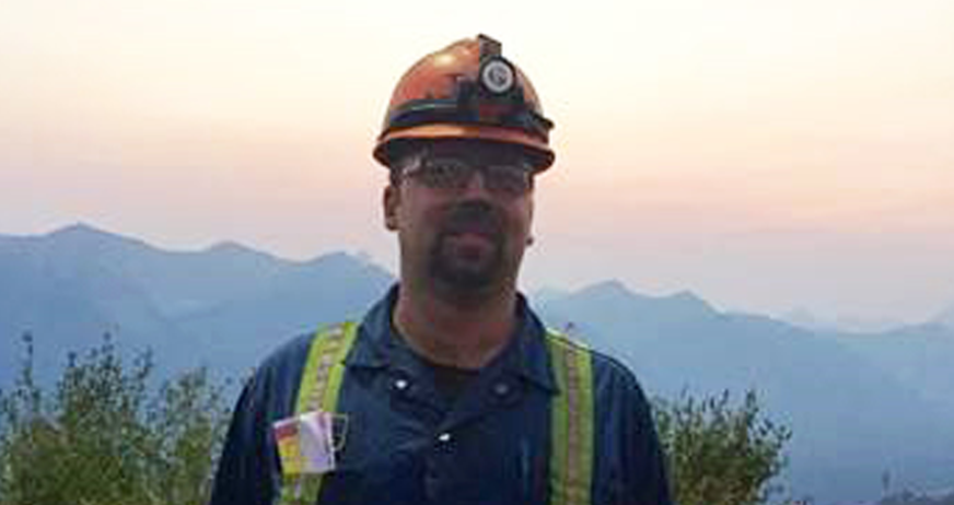 A man in a hard hat and safety gear standing outside with mountains and hazy sky in the background.